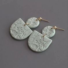 two pairs of white ceramic earrings with gold ear wires on a gray surface, one is shaped like an oval and the other has leaves