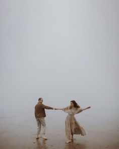 a man and woman holding hands while standing in the sand on a foggy day