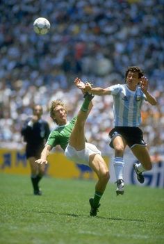 two men playing soccer on a field with people watching from the stands in the background