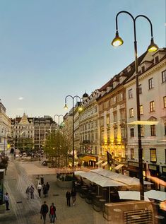 people are walking around in the city at sunset or sunrise time with street lamps and buildings on either side