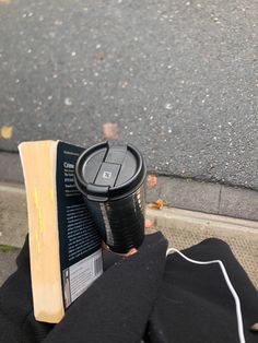 a person is holding a book and a coffee cup on their lap while sitting on the curb