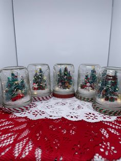 four glass jars with christmas trees in them on a red doily tablecloth and white lace