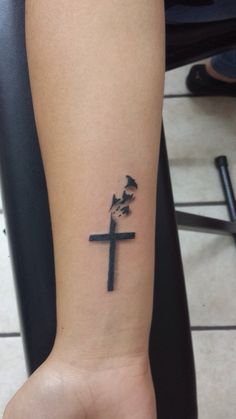 a small cross tattoo on the left inner arm, with an inked dove above it