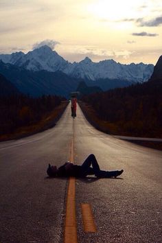 a person laying on the side of an empty road with mountains in the background at sunset