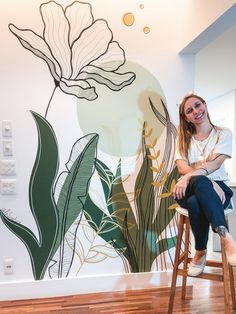 a woman sitting on a stool in front of a wall with flowers painted on it