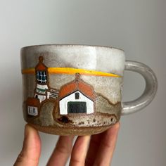 a hand holding a coffee cup with a painting on the side and a small house painted on it