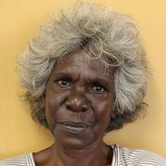 an older woman with blonde hair and blue eyes looks at the camera while standing against a yellow wall