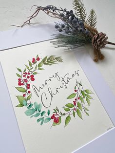 a christmas card with watercolor leaves and berries on it next to a pine cone