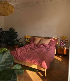 a bed with pink sheets and pillows in a room next to potted plants on the floor