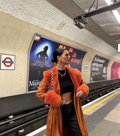 a woman in an orange coat and black top standing on a subway platform