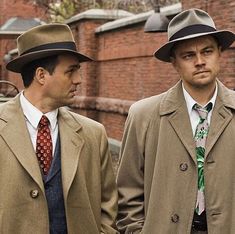 two men in coats and ties standing next to each other near a building with brick walls