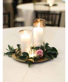 three candles are lit on a gold plate with greenery and leaves around it, surrounded by white tablecloths