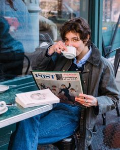 a young man sitting at a table drinking from a coffee cup and reading a magazine