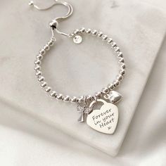 Our beloved memorial bracelet designed to commemorate cherished memories - now features a sweet angel charm This beautiful silver bracelet combines a silver heart charm which can be engraved with a loved one's name, alongside a pretty silver angel and a dainty silver heart charm. The sterling silver heart charm measures 16mm across the front of the heart, perfect for engraving a name or a message, and sits centrally on a sterling silver ball slider bracelet crafted with 4mm silver beads. With a Sterling Silver Name Bracelet With Heart Charm For Gift, Silver Name Bracelet With Heart Charm For Gift, Silver Name Bracelet With Heart Charm As Gift, Silver Name Bracelet With Heart Charm, Silver Sterling Name Bracelet With Heart Charm, Sterling Silver Heart Name Bracelet For Anniversary, Personalized Sterling Silver Bracelet With Heart Charm, Engraved Sterling Silver Charm Bracelet For Mother's Day, White Gold Charm Bracelet For Anniversary On Mother's Day