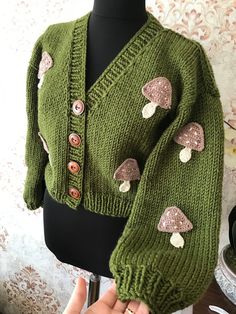 a green knitted sweater with mushrooms on the sleeves and buttons, being held up by a mannequin