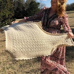 a woman is holding up a crocheted shawl in the grass with trees in the background