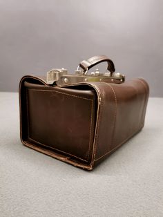 The brown leather bag from the 1960s exudes vintage charm and functionality. This elegant bag likely served doctors or veterinarians of that era to carry their precious tools. With its triangular profile and sturdy leather handles, the bag appeared serious and authoritative. The bag's interior was probably lined with soft material to protect medical or veterinary instruments. This well-designed bag was not only practical but also reflected the professional reputation of its owner. Over time, this bag has become a valuable collector's item. Its quality craftsmanship and authentic vintage appearance make it special. Whether used as a functional piece or as a decorative element in an interior, the brown leather bag from the 1960s leaves a strong impression and carries with it a story of its m Retro Leather Box Bag For Travel, Brown Satchel With Palladium Hardware For Formal Occasions, Retro Leather Box Bag With Top Handle, Retro Leather Top Handle Box Bag, Retro Leather Satchel Box Bag, Vintage Rectangular Leather Box Bag, Vintage Brown Satchel With Palladium Hardware, Classic Brown Box Bag With Handle Drop, Vintage Shoulder Bag With Palladium Hardware For Business