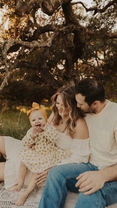 a man, woman and baby sitting on a blanket in front of a large tree