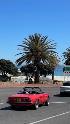 a red convertible car driving down a street next to a palm tree and ocean in the background