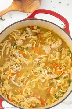 a red pot filled with chicken noodle soup on top of a white counter next to a wooden spoon