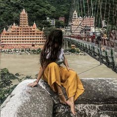 a woman sitting on top of a stone wall next to a suspension bridge over a river