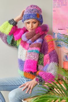 a woman wearing a multicolored sweater and hat sits on a stool in front of a potted plant