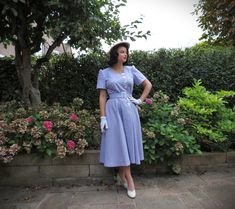 a woman in a blue dress and white gloves standing next to some bushes with pink flowers