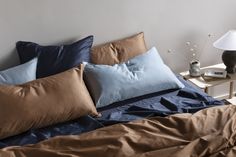 an unmade bed with blue and brown pillows on it, next to a night stand