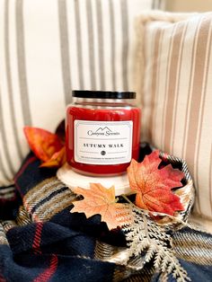 a jar of autumn wax sitting on top of a plaid blanket next to an orange maple leaf