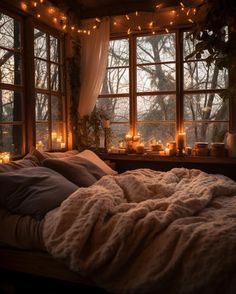 an unmade bed in front of two large windows with candles on the window sill