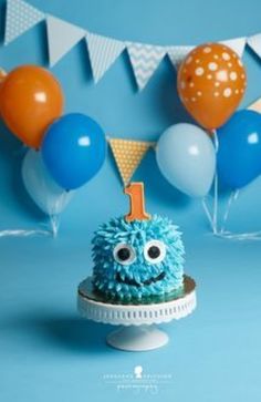 a blue cake with an one on top and balloons in the background for a 1st birthday party