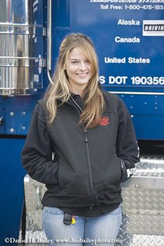 a woman standing in front of a blue semi truck smiling at the camera with her hands on her hips