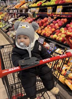 a baby sitting in a shopping cart at a grocery store wearing a hat and pacifier