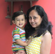 a woman holding a little boy in her arms and smiling at the camera while standing next to a red wall