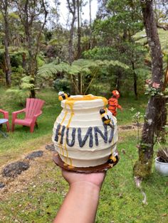 a hand holding up a ceramic jar with the word humong written on it in front of some chairs and trees