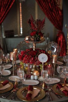 the table is set with plates, silverware and red flower centerpieces on it