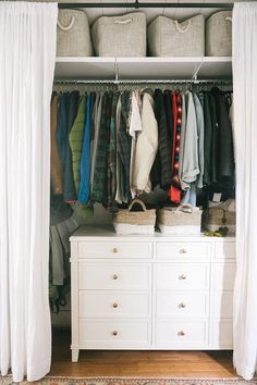 an organized closet with hanging clothes and baskets on the shelves, along with white curtains