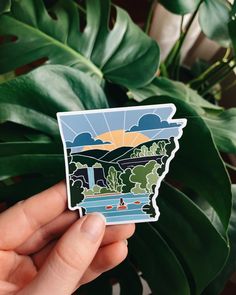 a person holding up a sticker with the image of a lake and mountains on it