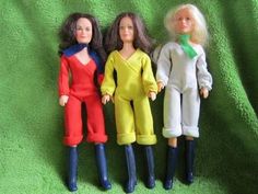 three dolls are standing next to each other on a green cloth covered surface, one is wearing blue boots and the other has red pants