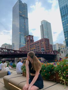 a woman sitting on top of a bench in front of tall buildings and people eating