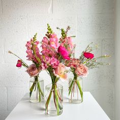 three glass vases with pink flowers in them on a white countertop next to a brick wall