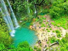 people standing in front of a waterfall with blue water and green trees around it, surrounded by lush greenery