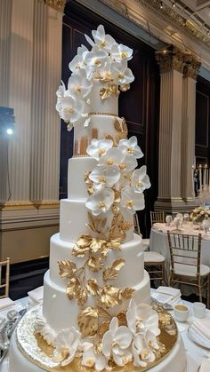 a tall white and gold wedding cake with flowers on it's side at a banquet table