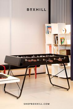 A black outdoor football table with wooden handles set in a minimalist indoor space with white shelving unit filled with books