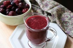 a glass mug filled with red liquid next to a bowl of cherries on a white plate