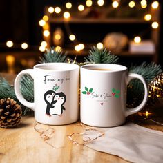 two coffee mugs with penguins on them sitting next to pine cones and christmas lights