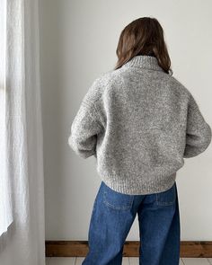 a woman standing in front of a window with her back to the camera, wearing jeans and a sweater