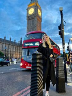 a woman standing on the sidewalk in front of a clock tower with a red double decker bus behind her