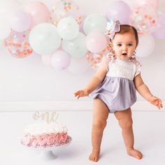 a baby girl standing in front of a cake with balloons behind her and a first birthday cake