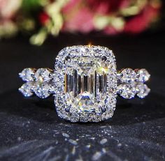 an engagement ring with a large diamond surrounded by smaller diamonds on a black surface and flowers in the background